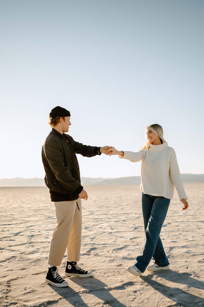 Couple dances on the Bonneville Salt Flats in Utah. The couple is demonstrating an example of what to wear for your engagement photoshoot.  He is wearing black converse shoes, tan pants, black bomber jacket, and a black beanie. She is wearing white sneakers, blue jeans, cream sweater. 