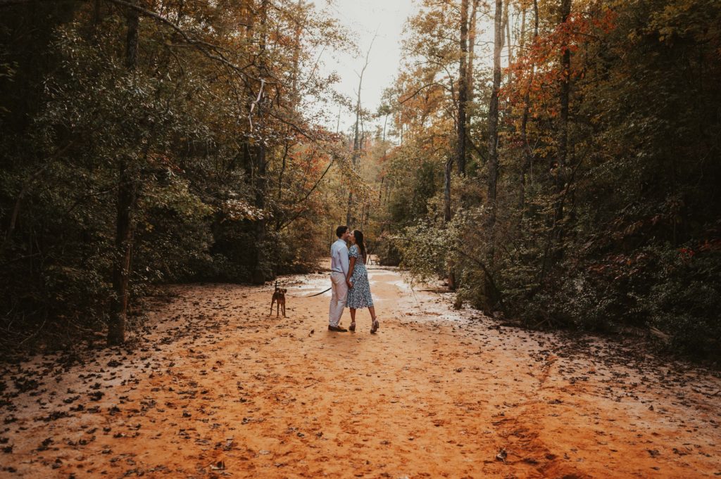 Couple standing in the middle of the frame facing each other and about to go in for a kiss. They are holding their dog on a leash. Standing in a red soil canyon with trees surrounding them with fall colors.