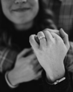 Black & White image of an engaged couple shortly after a surprise proposal. A woman is behind hugged from behind by her partner and they are showing off the new ring.