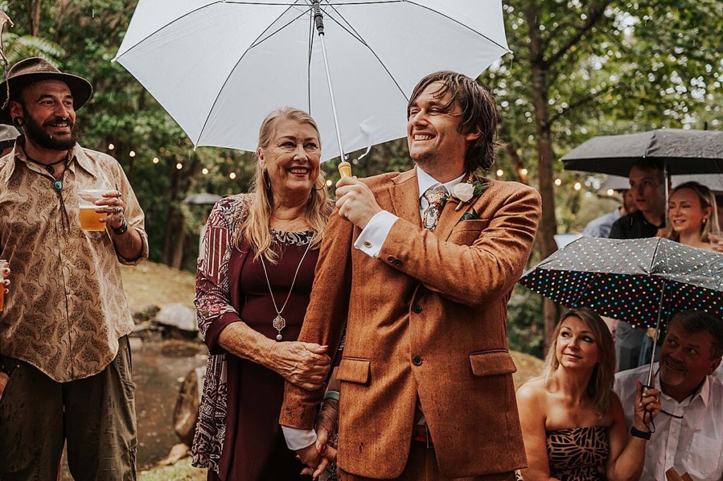 Groom in a burnt orange suit walking down the aise with his mom and an umbrella