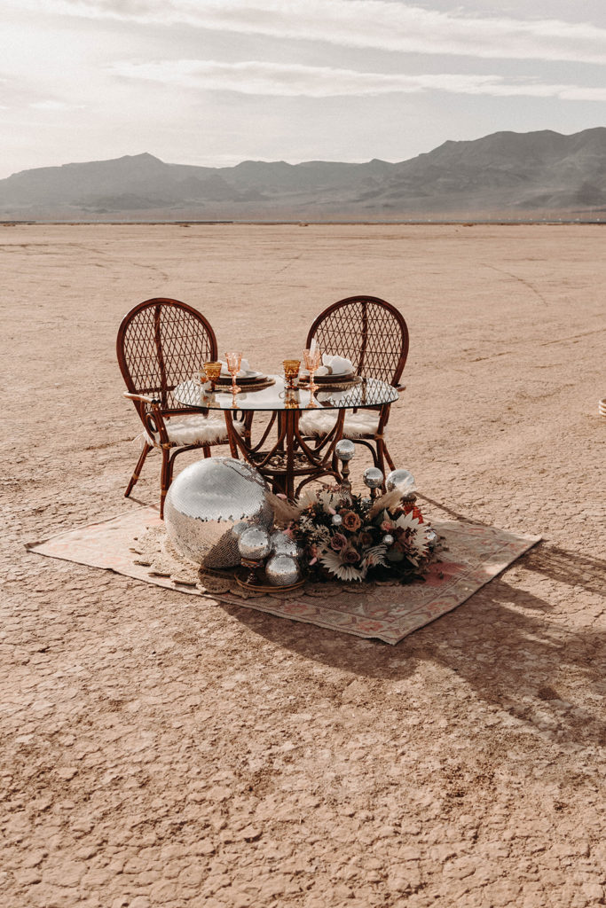 Boho desert elopement sweetheart table with rattan chairs, disco balls, a rug and florals.
