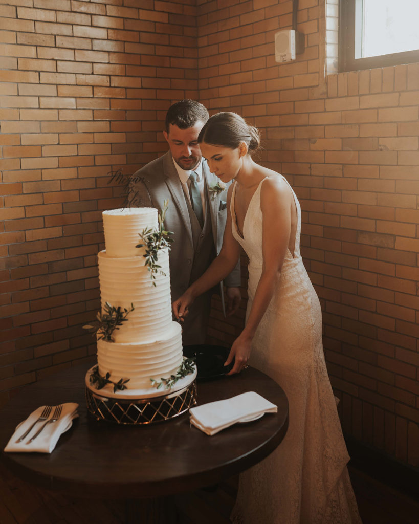 Bride and groom cutting their 3-tier white buttercream wedding cake with greenery. 