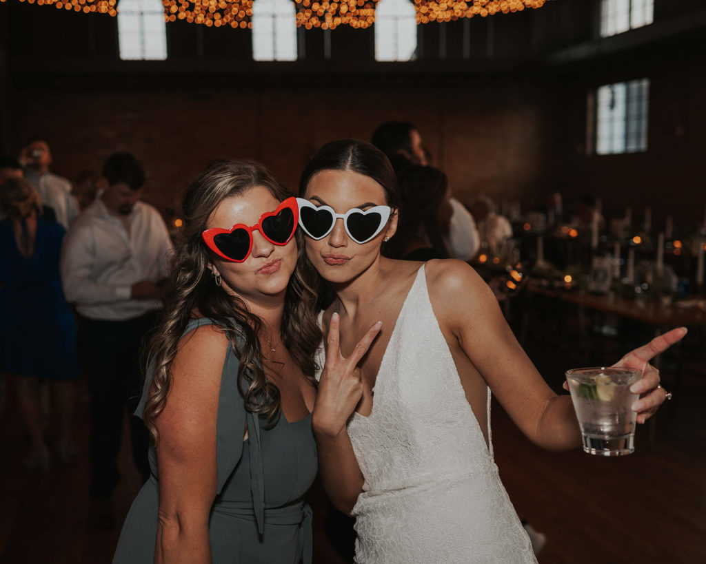 Bride and bridesmaid wearing heart-shaped glasses at the reception.