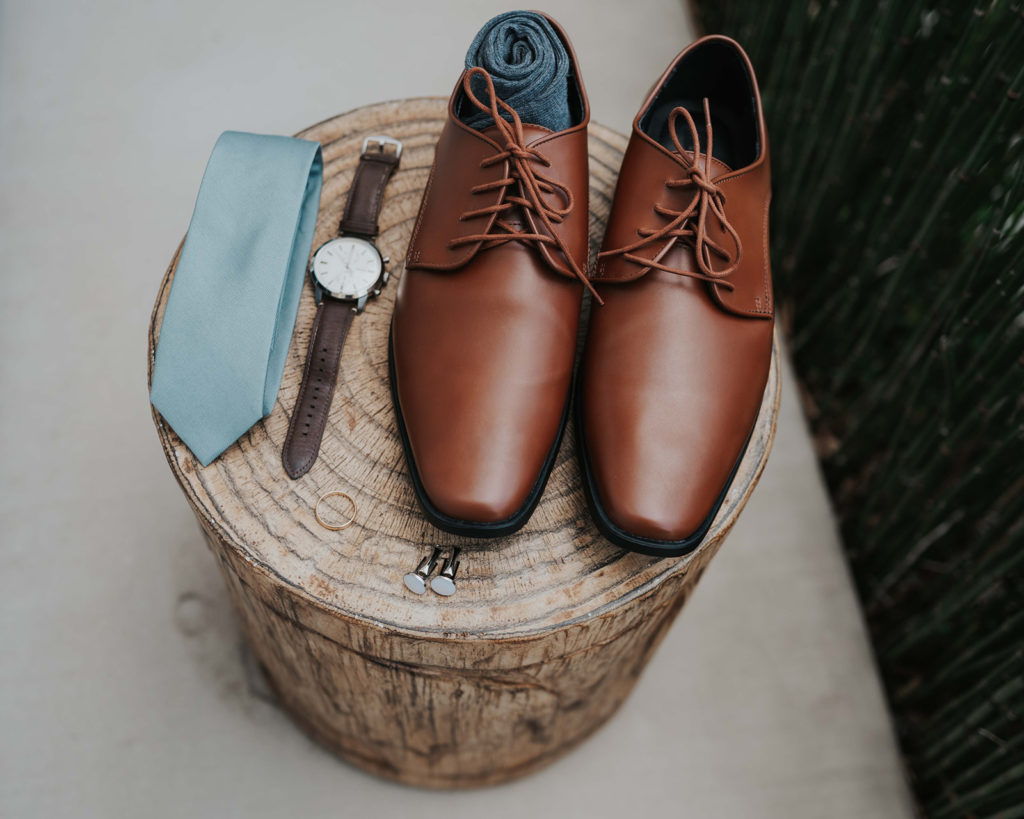 Groom wedding detail flat lay featuring his brown dress shoes, rolled up socks, blue tie, cuff links and watch on a block of wood.