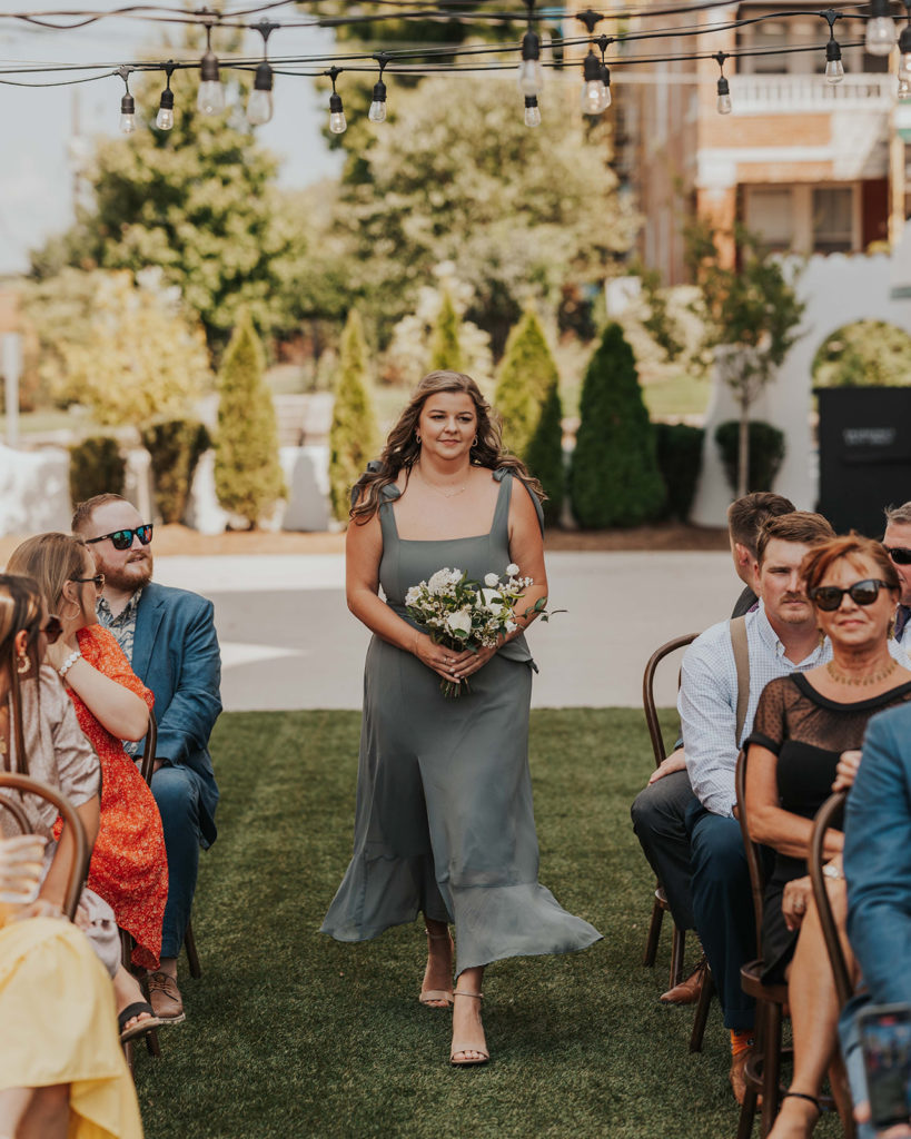 Bridesmaid in dusty blue dress walking down the aisle at this outdoor ceremony at the Common House in Chattanooga, TN.