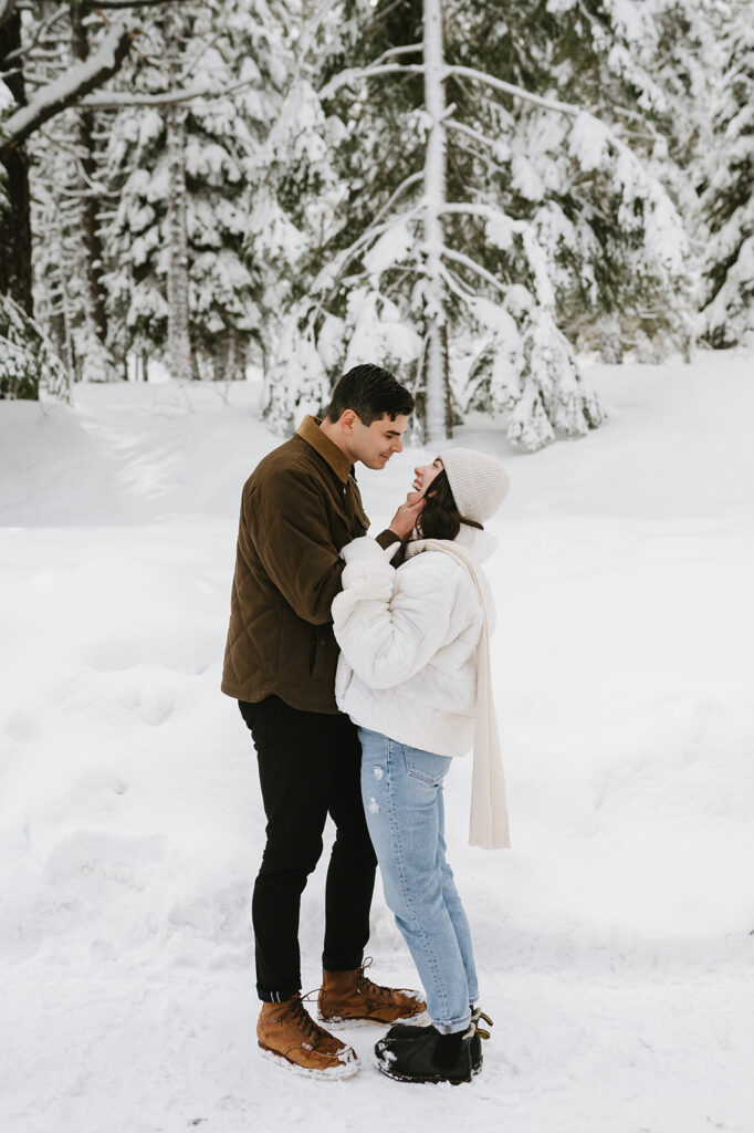 Couple stands in snowy forest during their engagement photos by Sarah Woods.