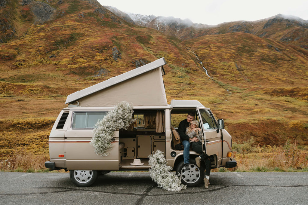 A vintage camper van is parked in front of foggy mountains in the Fall. The photoshoot location is Hatcher's Pass, Alaska. Large installations of Baby's Breath flowers appear to be pouring out of the van. A couple sits in the passenger seat with the door open. They both have their legs out the door and the woman is sitting in between her husband's legs.
