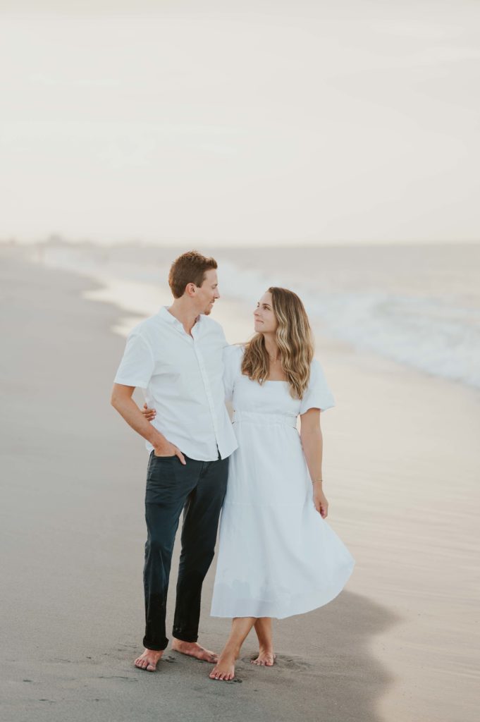 Engagement photographs on Melbourne Beach in Florida. The couple is demonstrating an example of what to wear for your engagement photoshoot. She is wearing a white long dress and he is wearing a white button up and navy chinos. 

