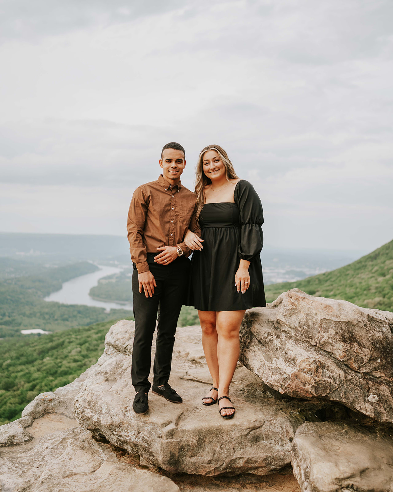 Couple stands on rock with Chattanooga Tennessee in the background. They are demonstrating an example of what to wear for your engagement photoshoot. Man is wearing brown button up, with black pants and black dress shoes. His wife is wearing a black long sleeve dress with sandals.