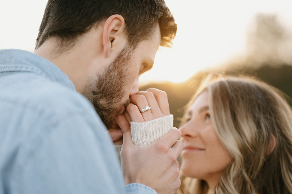 Man kisses woman's hands while both looking at each other. Photoshoot location is blurred. The sun is peeking through the background. On the woman's hands is an engagement ring.