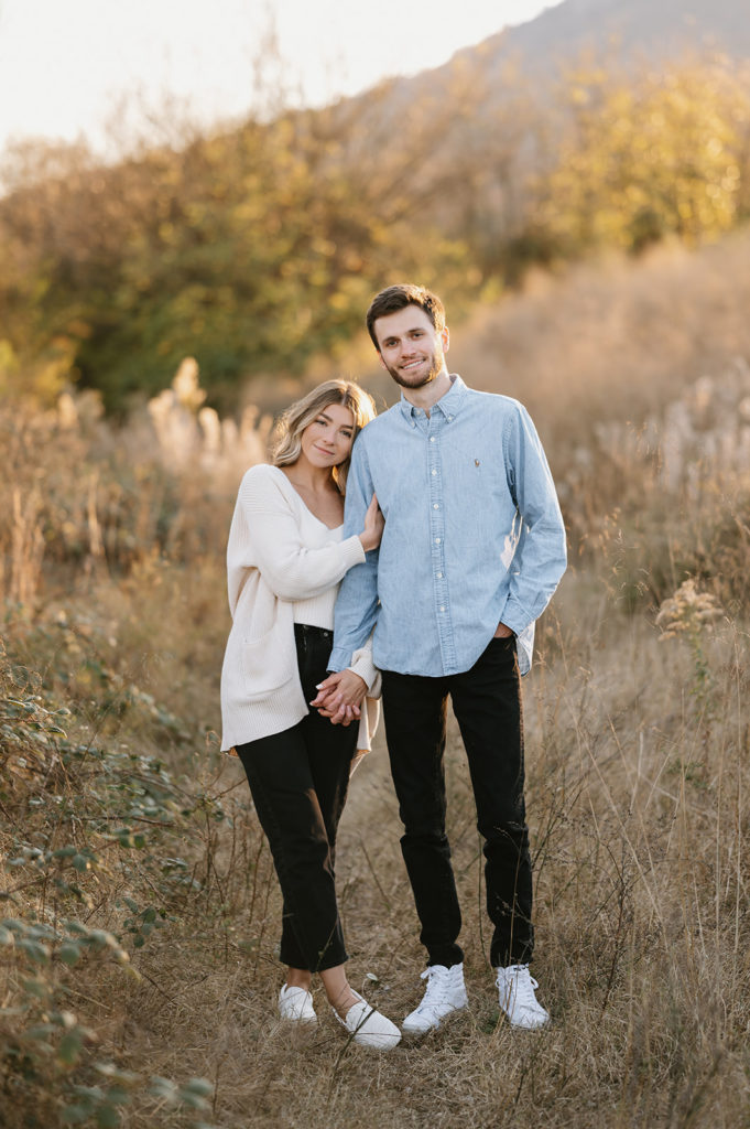 Couple is standing in a field. They are demonstrating an example of what to wear for your engagement photoshoot. They are both wearing black jeans. Girl is wearing a white tank top + cardigan. Boy is wearing blue button up. 