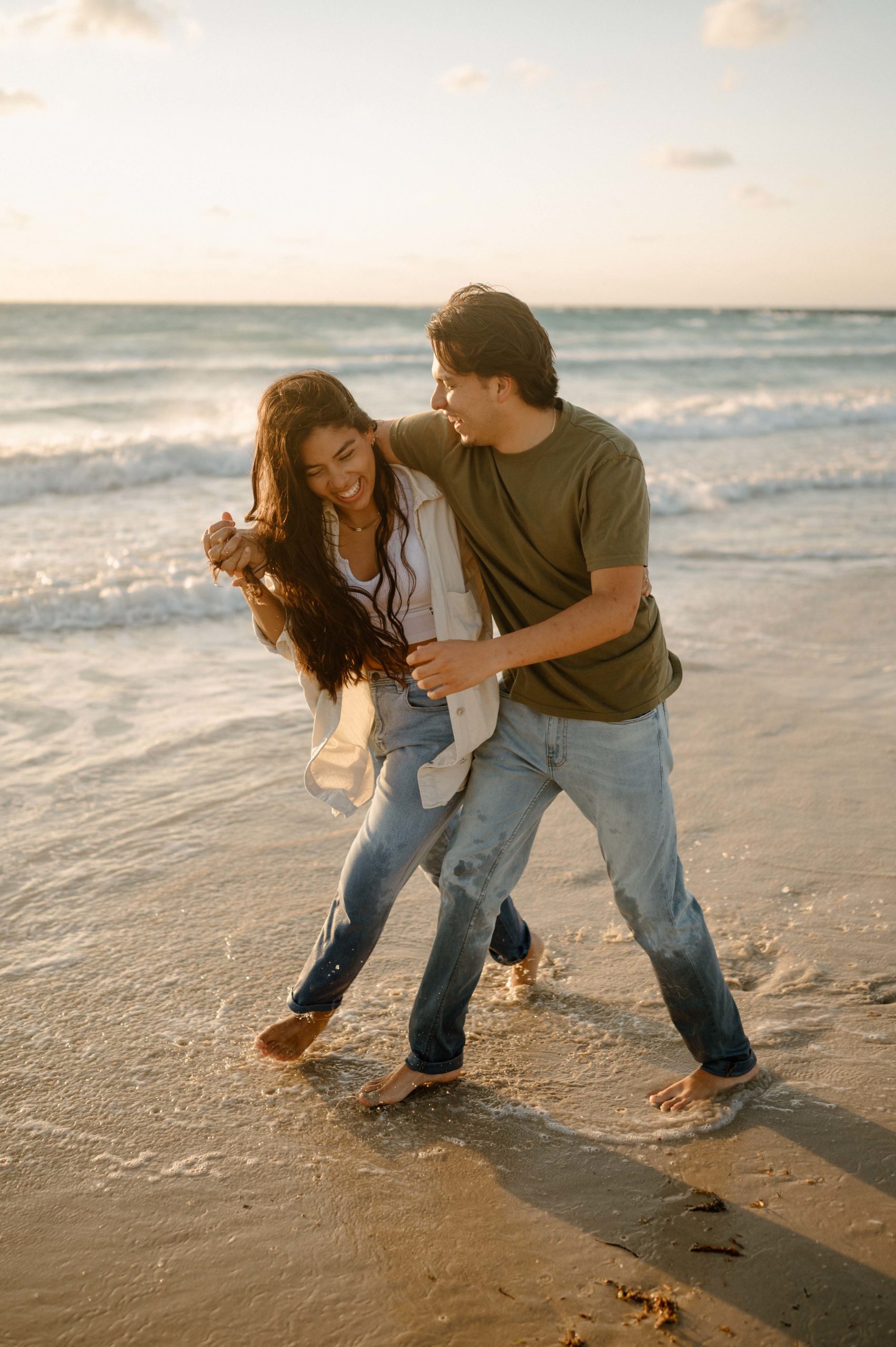 Couple walks down the beach during sunrise while laughing. They are demonstrating an example of what to wear for your engagement photoshoot. They are both wearing light jeans and neutral colored shirts 