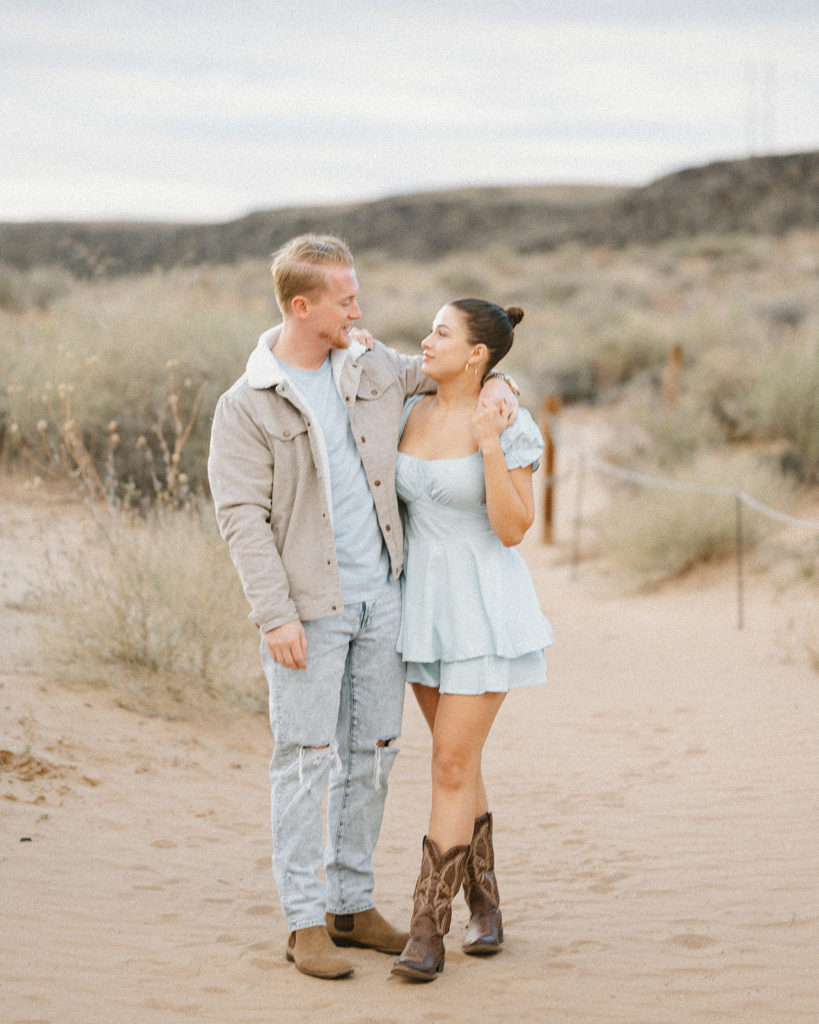 Couple stands in the desert. Girl is wearing light blue dress and brown boots. Man is wearing light blue jeans and light blue tshirt, with a tan jacket. They are demonstrating an example of what to wear for your engagement photoshoot.