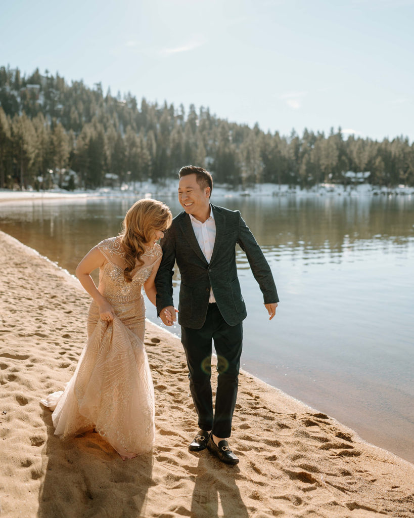 Husband and wife walk and laugh on the beach. The photoshoot location is in Lake Tahoe, Nevada. The are dressed in formal attire. The water is extremely calm. There is snowy evergreen trees in the background with blue skies.