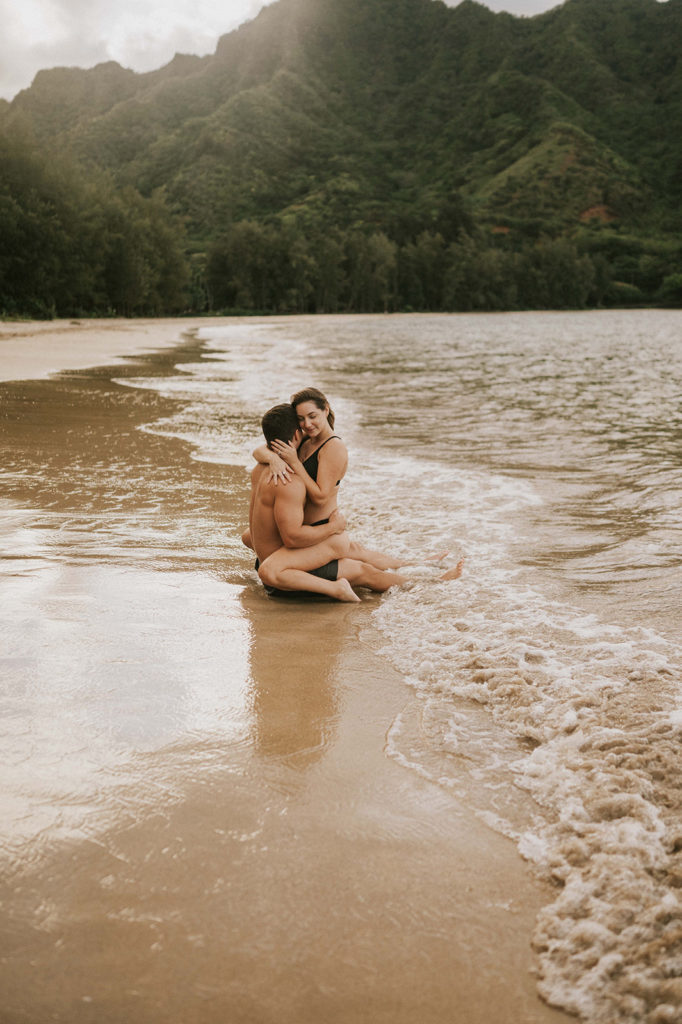 Husband and Wife are sitting on a beach. The photoshoot location is in Oahu, Hawaii. The sun is setting behind them. She is sitting in his lap and facing towards him while wrapping him up for a hug. The ocean water is calm, and laps over their legs as the sit in the water.