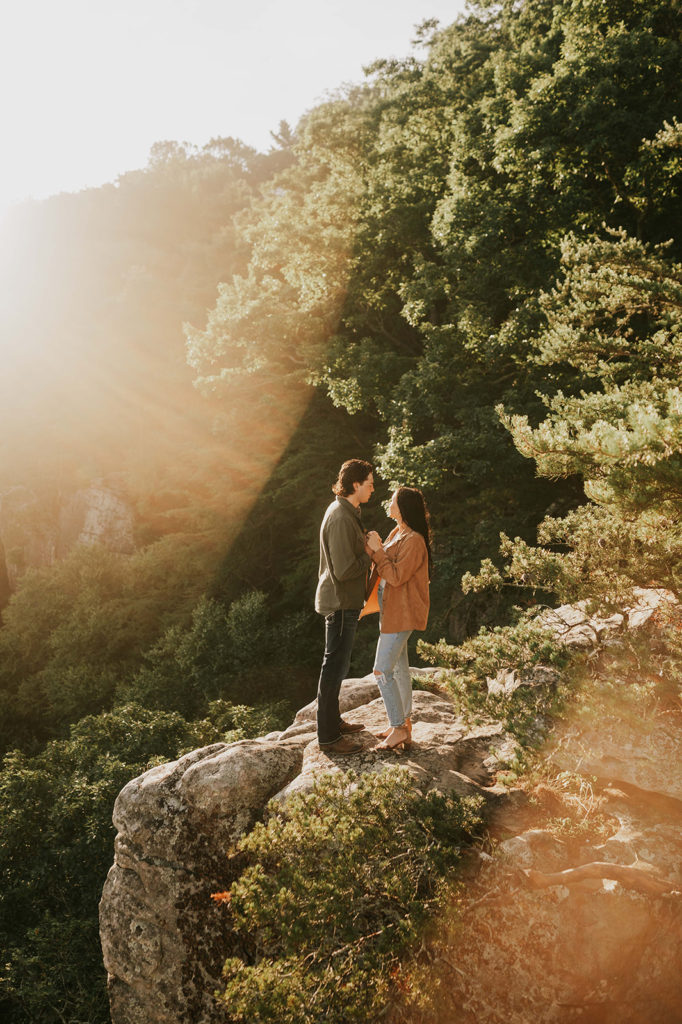 Man and Woman stand facing each other on a rocky cliff. The photoshoot location is in Chattanooga, Tennessee. They are holding hands and smiling at each other. The sunrise is coming up behind them.
