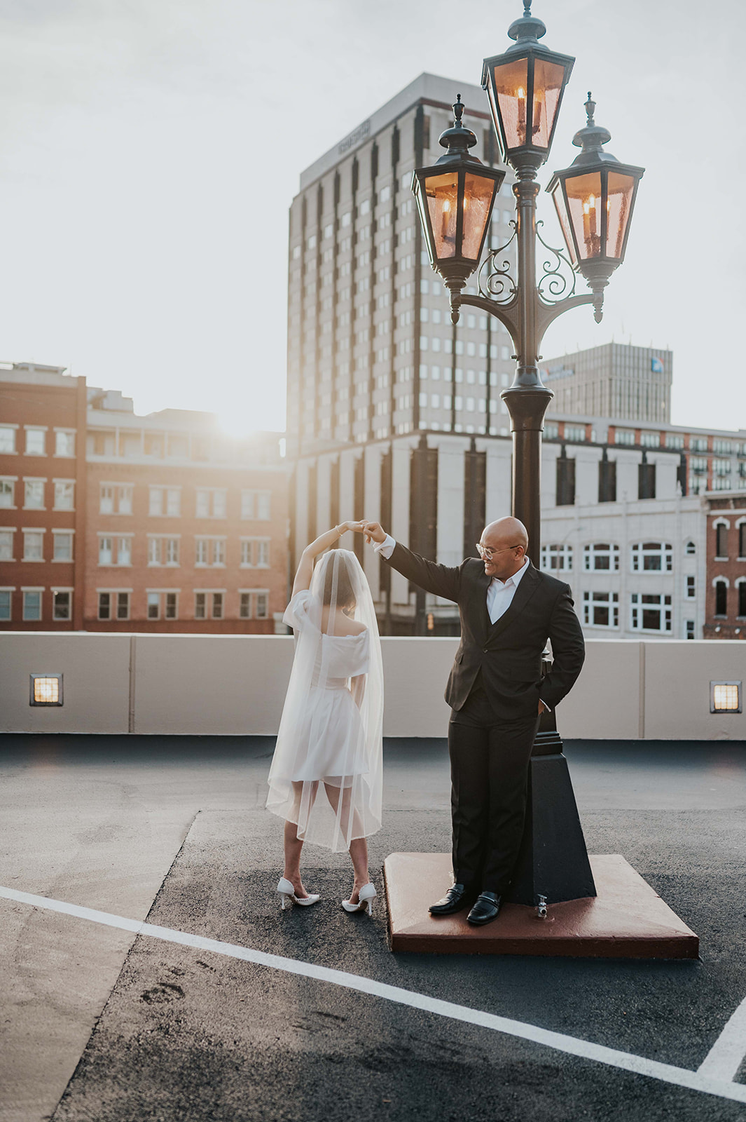 Downtown Engagement Photographs. Man is twirling his fiancé while they dance on a parking rooftop. They are demonstrating an example of what to wear for your engagement photoshoot. Man is wearing a black suit and woman is wearing a white dress and heels. 