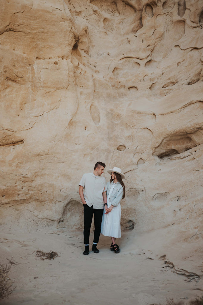 Couple stands in front of a tan rock face. They are demonstrating an example of what to wear for your engagement photoshoot. She is wearing a long white dress with a tan wide-brimmed hat. He is wearing black jeans and a tan shirt. 
