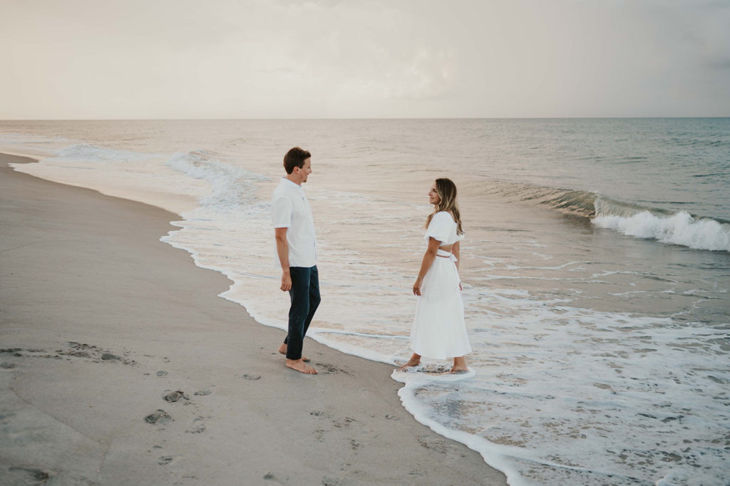 Man and woman smile at each other while standing on the beach during their beach engagement photographs.