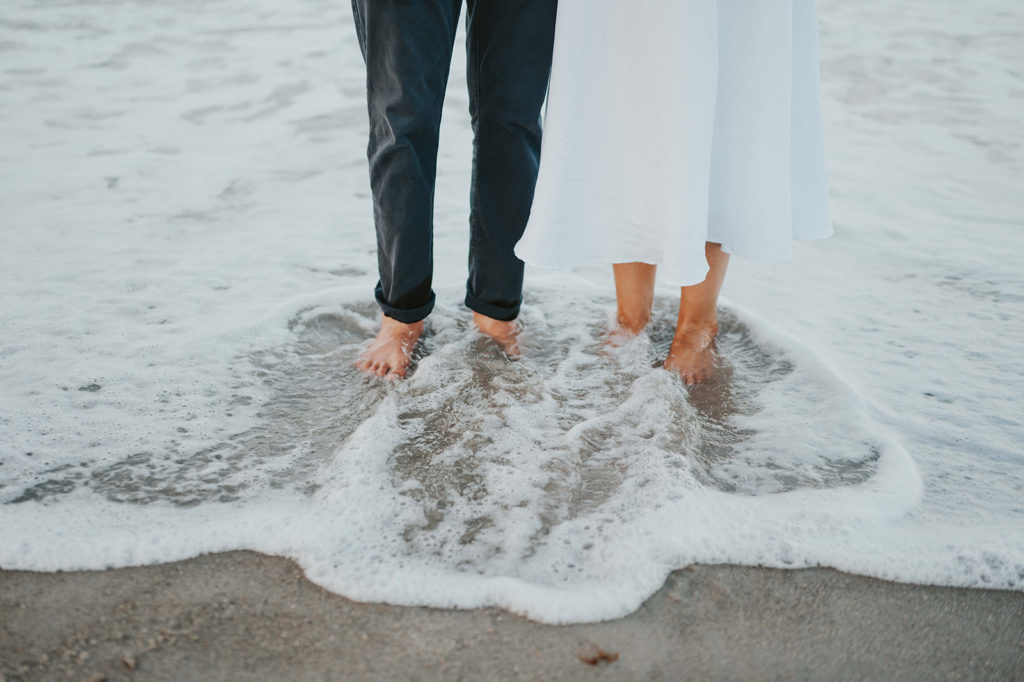 woman and man stand next to each other in the ocean shore line as the water flows over their feet 