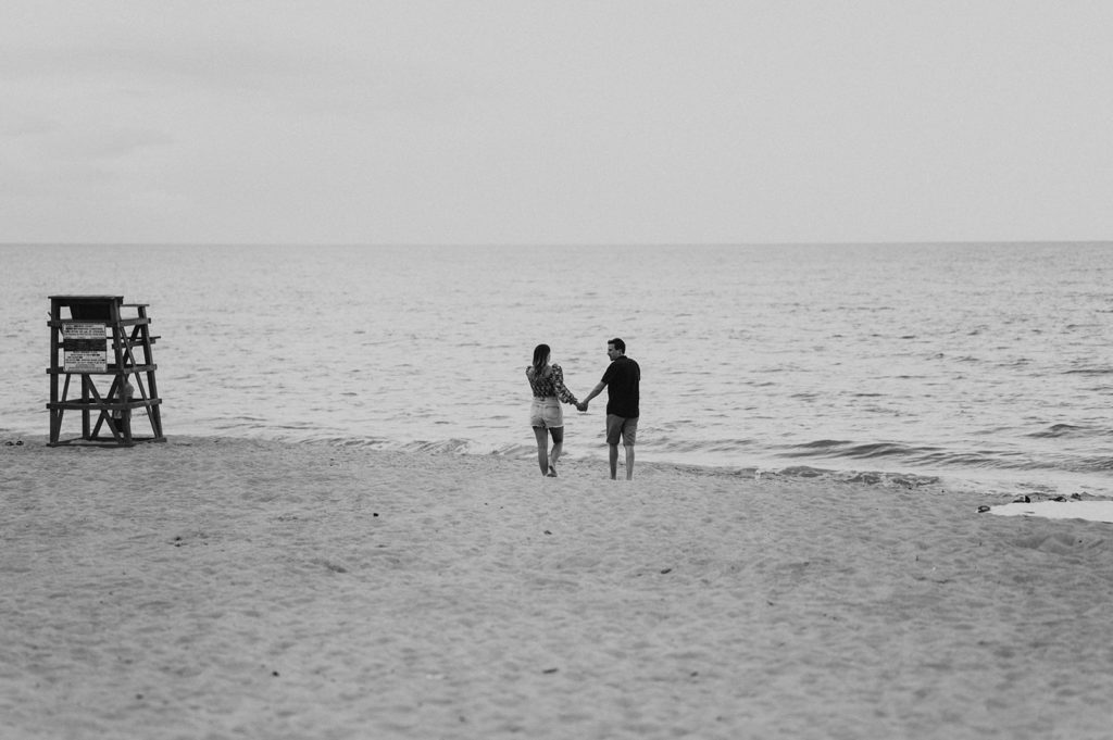 man and woman hold hands and walk towards the ocean during their beach engagement photos 