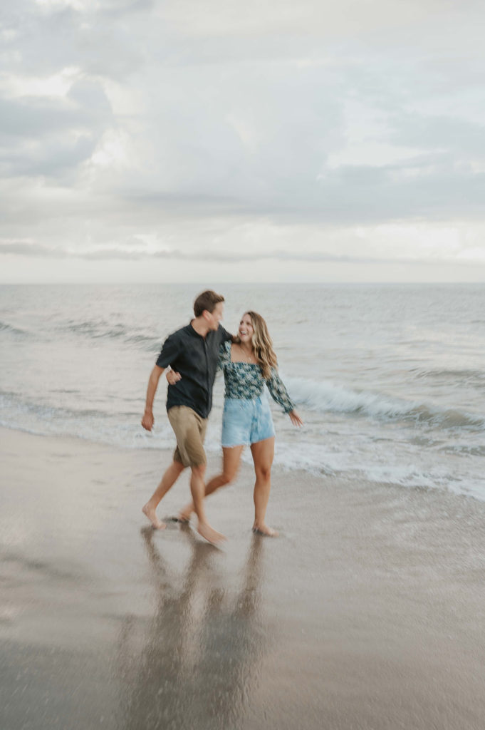 Man and woman walk along the ocean shoreline as they wrap their arms around each other during their beach engagement photos 