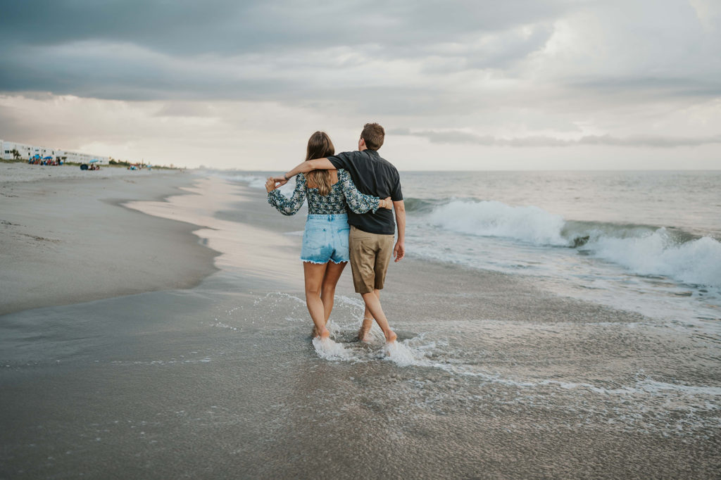 Man and woman walk along the ocean shoreline as they wrap their arms around each other 