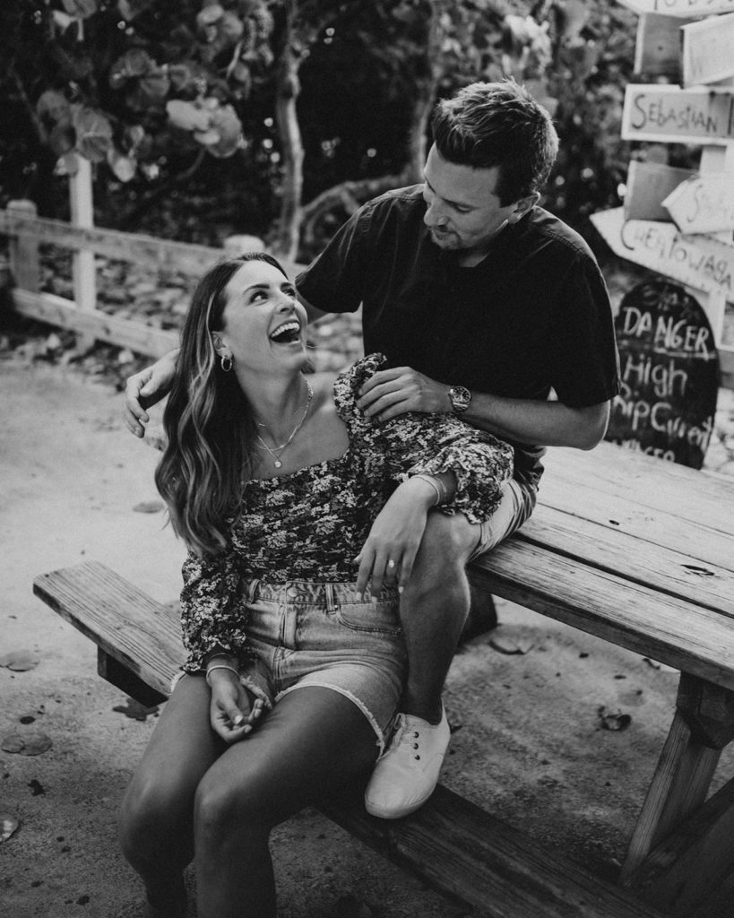 Man and woman sit on a picnic bench with the beach behind them. They are laughing together