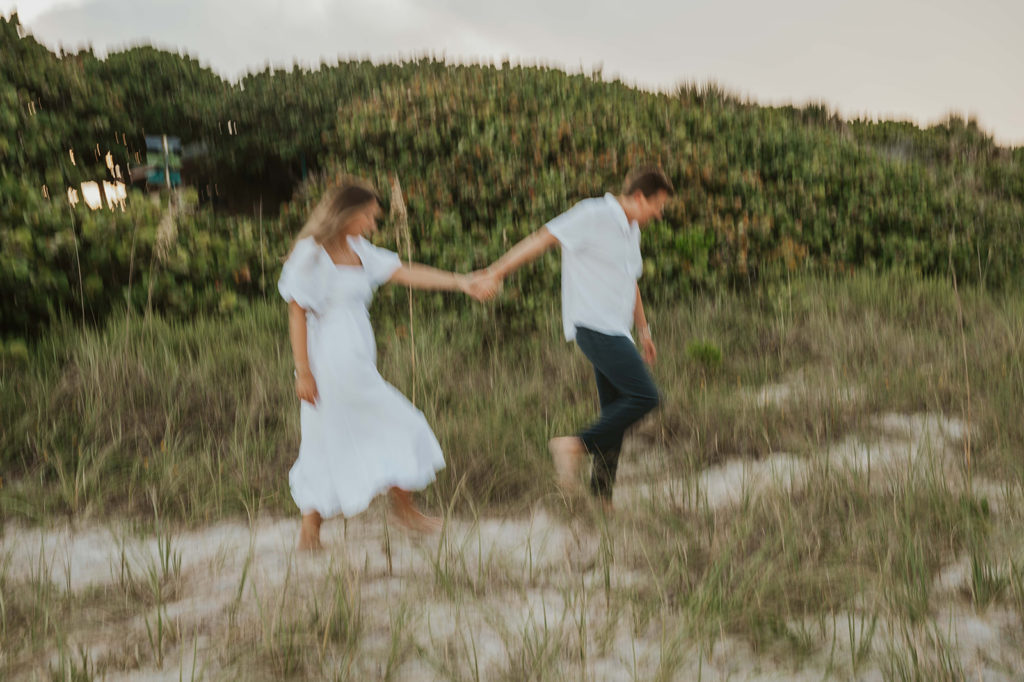 Man leads his fiancé through seagrass during their beach engagement photographs