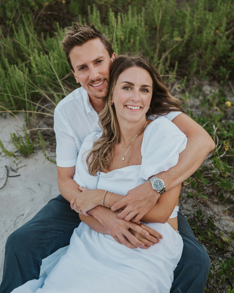 Couple is sitting on the ground during their beach engagement photographs. They are wrapped up in each other's arms