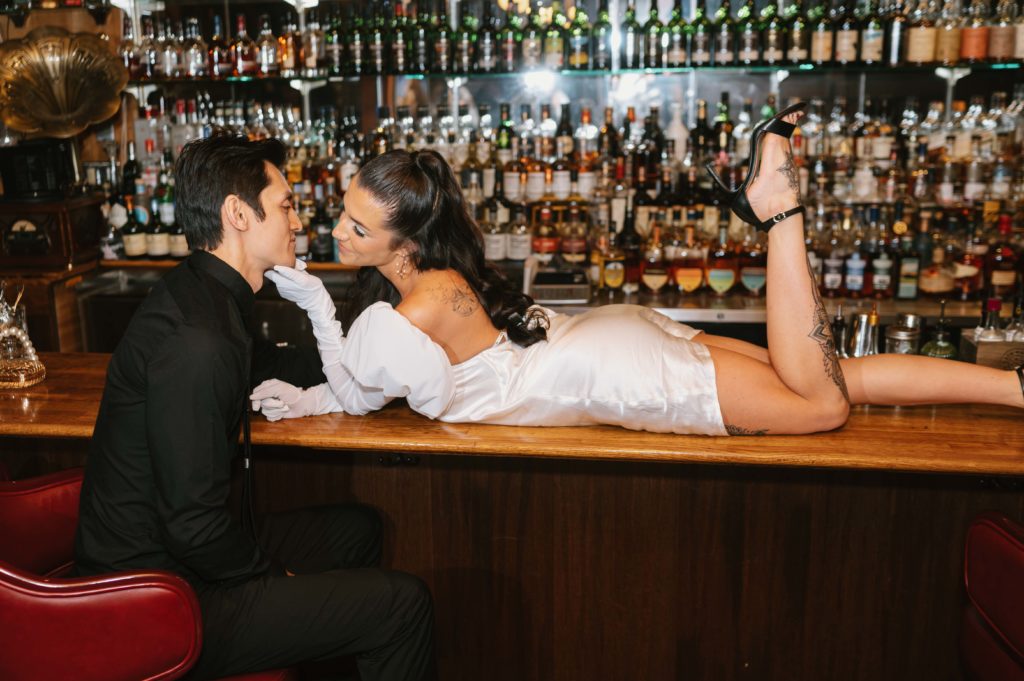 Bride lays on bar counter top and smiles at her groom. They are eloping at a local Italian restaurant.