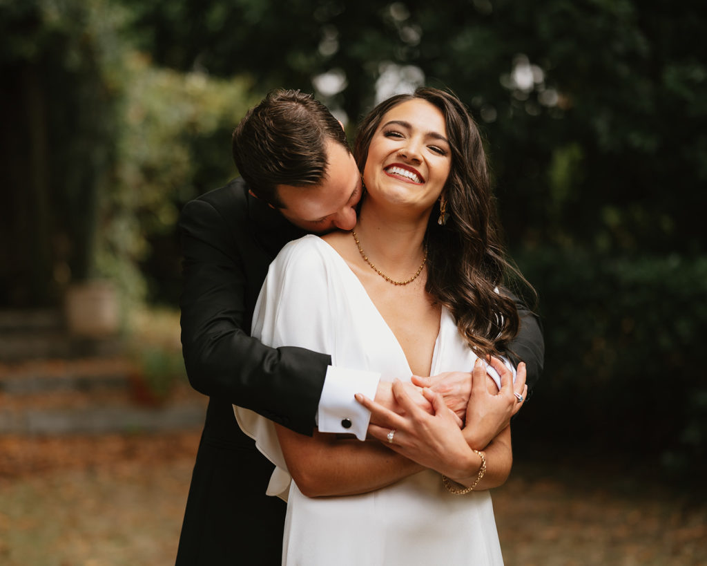 Groom hugs and kisses his Bride from behind. Bride smiles and wraps her arms around Groom.