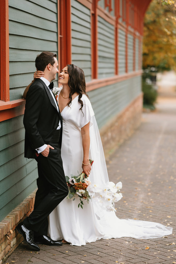 Bride and Groom are leaning up against a wall and smiling at each other
