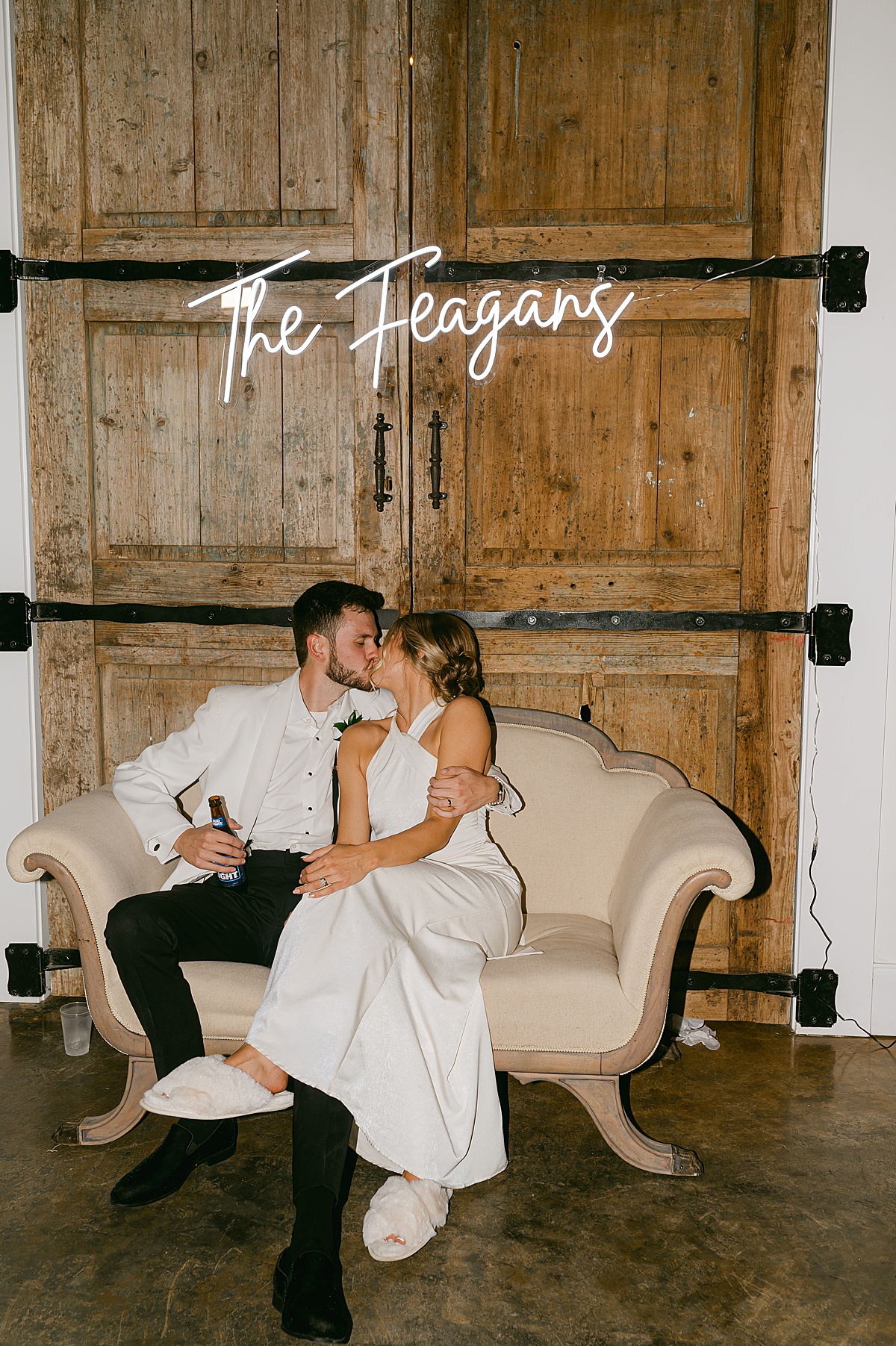Bride and groom sitting on a linen couch in front of a barn door at the Woodlands at Five Gables wedding venue kissing under their custom neon sign. 