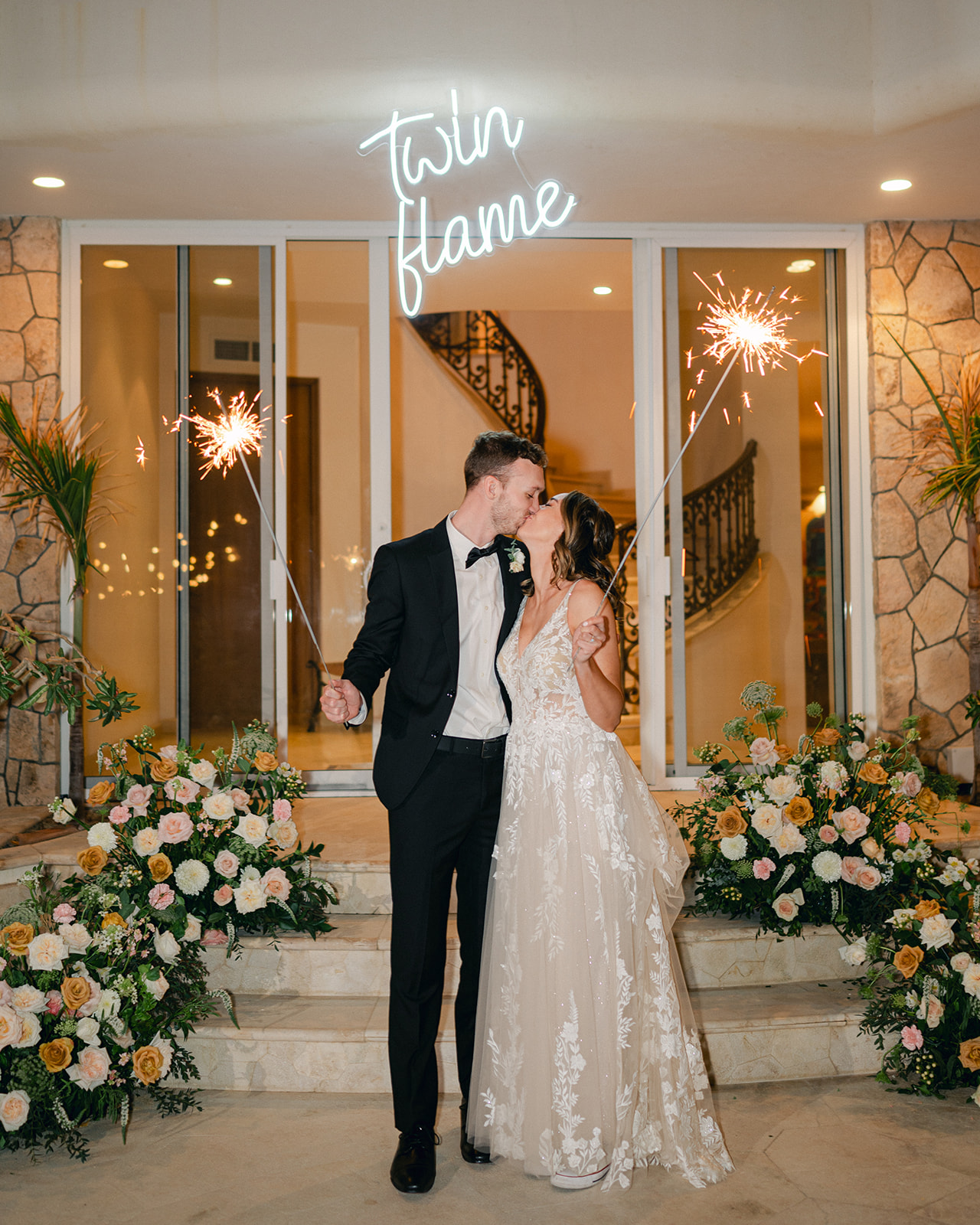 Bride and groom kissing under a neon "twin flame" sign holding sparklers. 