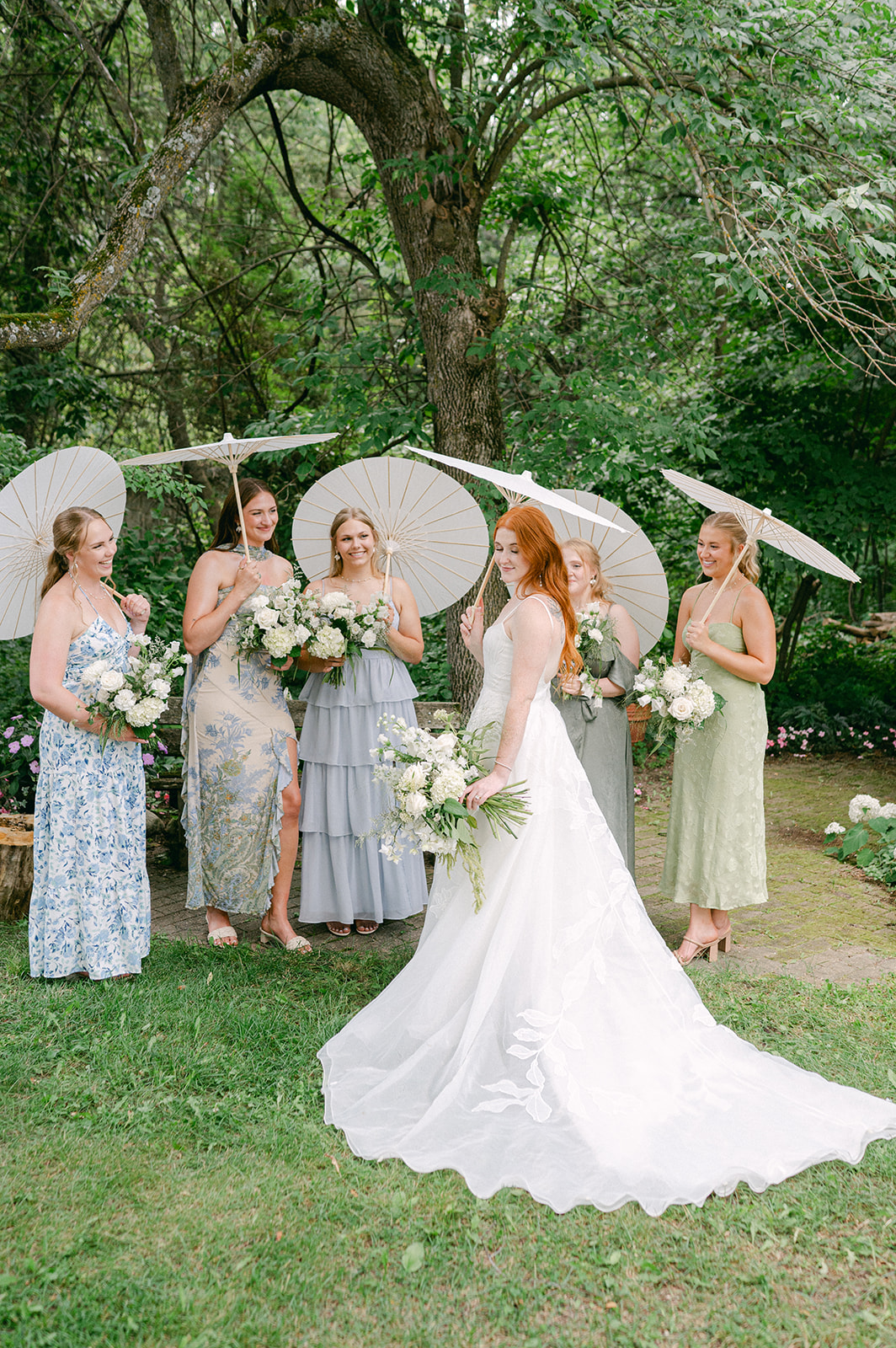 Cinematic garden wedding bridal party portrait, showcasing bridesmaids in blue and green floral dresses with white bouquets and parasols, set against natural greenery.