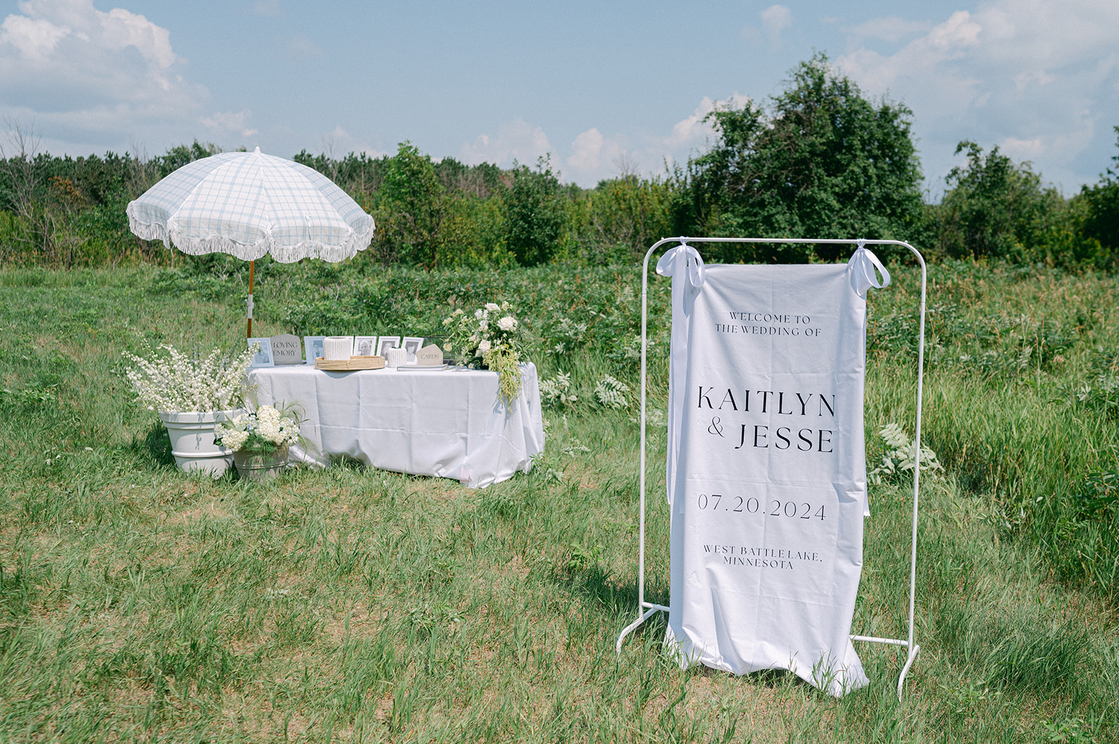 Garden party outdoor wedding ceremony welcome table with a white umbrella and fabric welcome sign. 