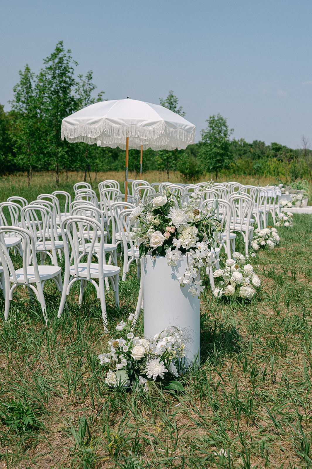 Outdoor summer ceremony in a field for a garden party wedding, featuring a white floral-lined aisle, elegant white chairs, and pedestals adorned with fresh blooms