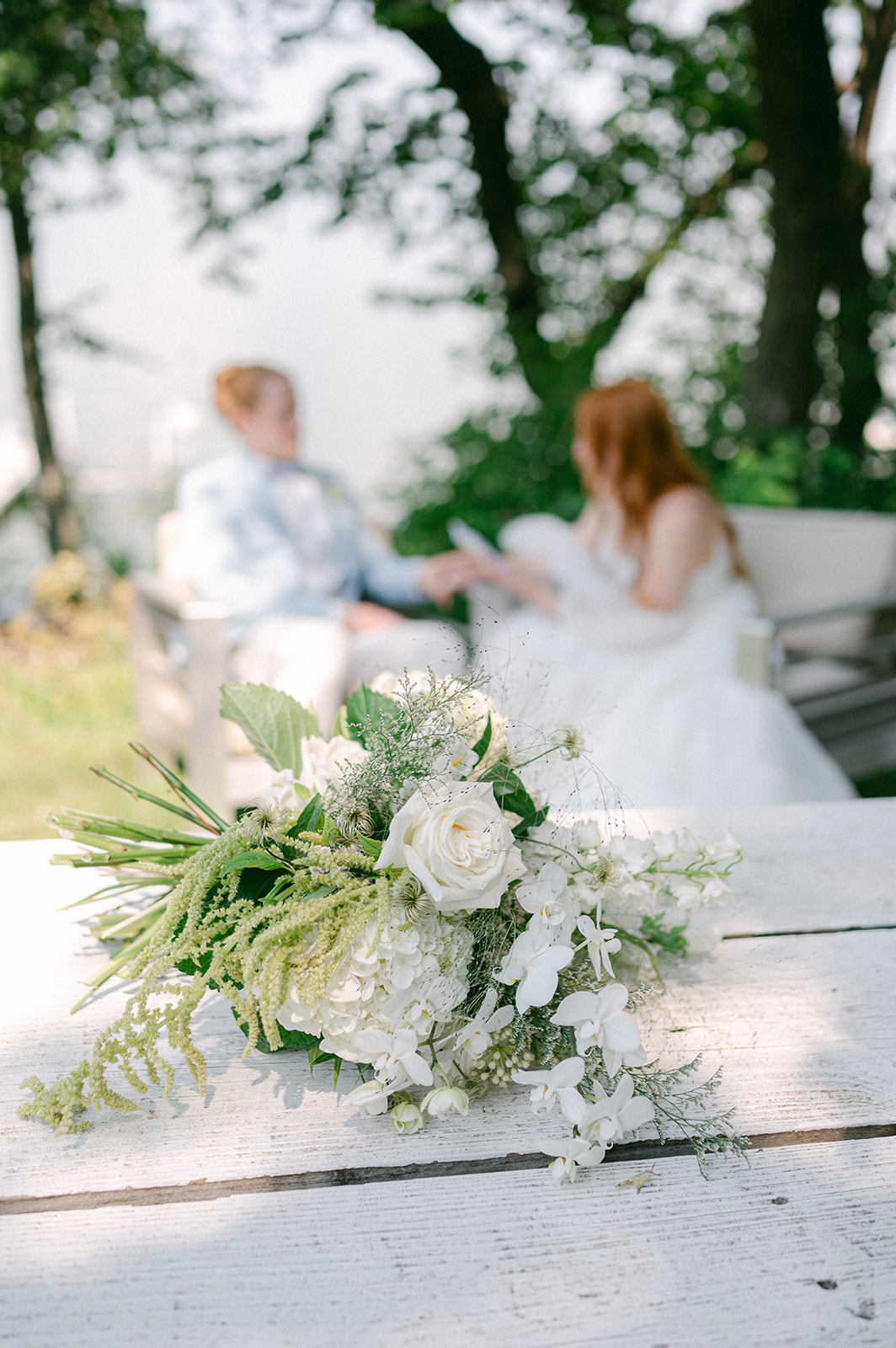 Bride's bouquet in focus while the couple shares a tender moment reading their vows in the background by the lake.