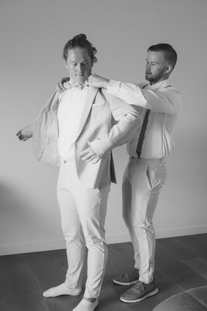 Black and white candid shot of the groom as his groomsman helps him put on his jacket.