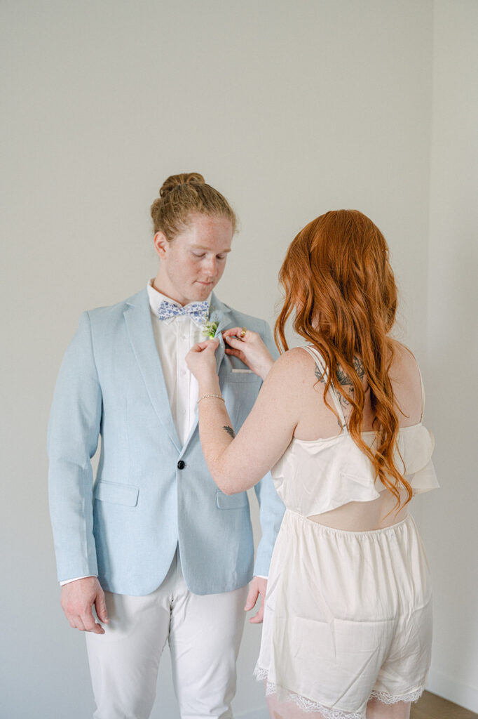 Bride in satin pajamas pinning the groom's boutonniere before their outdoor wedding.