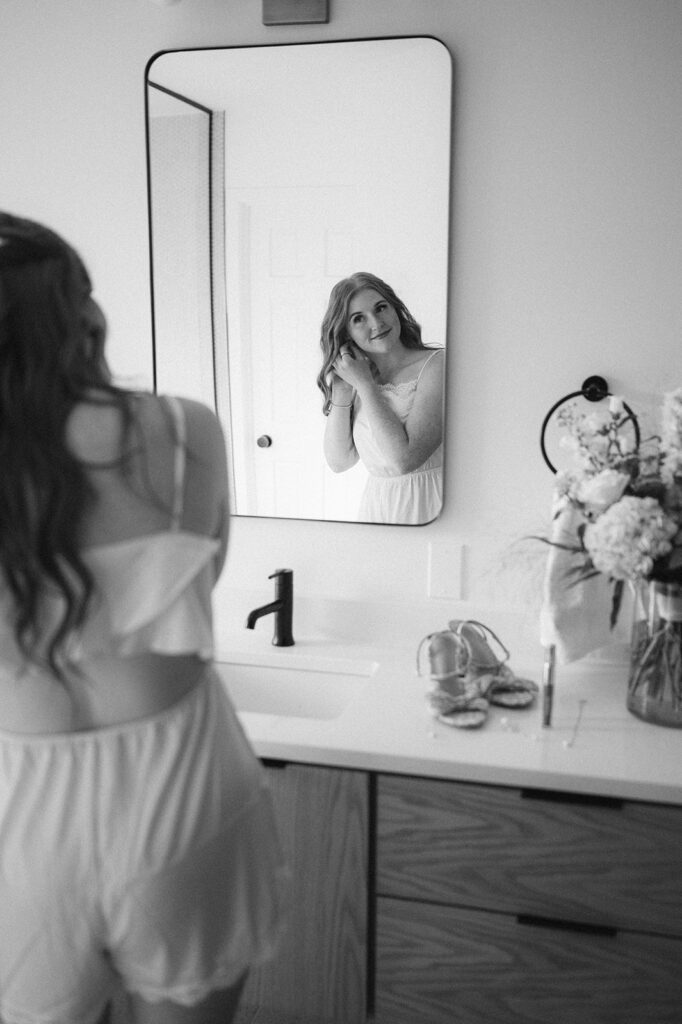 Black and white candid of bride in satin pajamas, captured through a mirror reflection while getting ready.