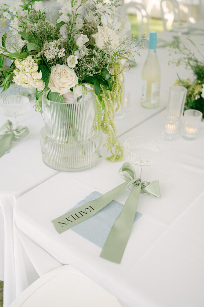 Wedding place settings adorned with green bows on personalized menus, white chairs, and lush floral centerpieces.