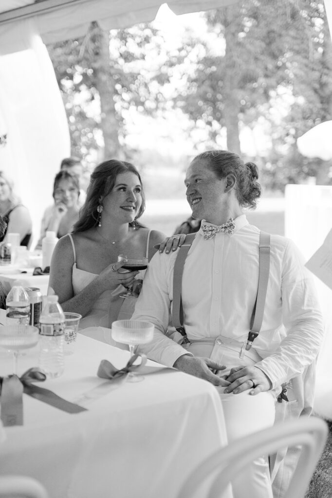 Candid shot of the bride and groom sitting together at their reception, smiling as they listen to heartfelt speeches from their loved ones.