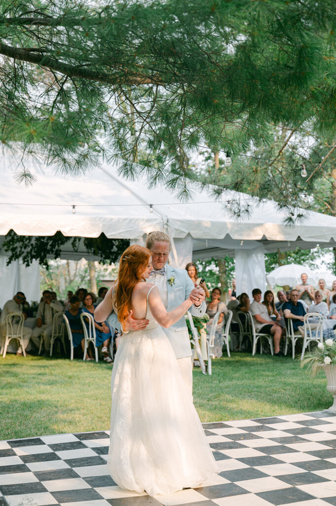 Candid moment of the bride and groom sharing their first dance on a checkered dance floor, surrounded by guests.