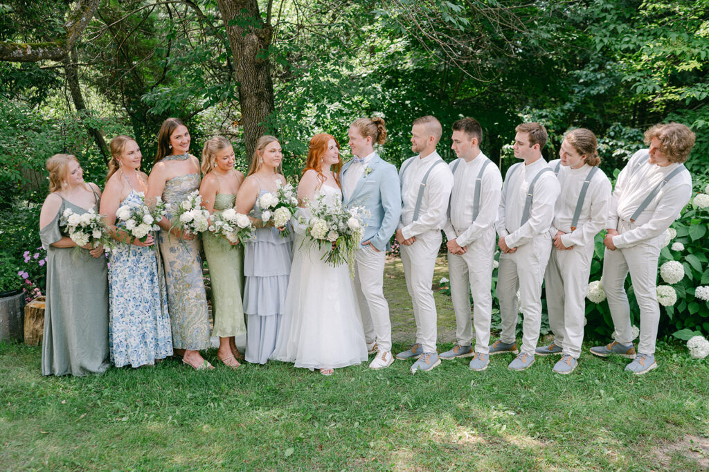 Portrait of the wedding party looking at the bride and groom in the garden, showcasing bridesmaids in blue and green floral dresses holding white bouquets.