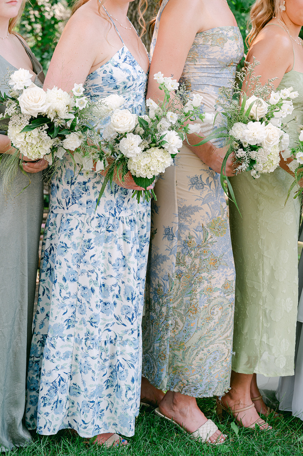 Bridesmaids in mismatched floral dresses in shades of blue and green with white bouquets.