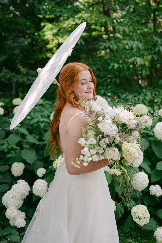 Editorial, fine art bridal portrait of a bride holding a vintage parasol umbrella and white bouquet in front of a hydrangea bush. 