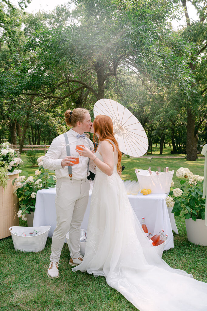 Bride and groom sharing a kiss while holding cocktails at their summer garden party wedding at Battle Lake. 