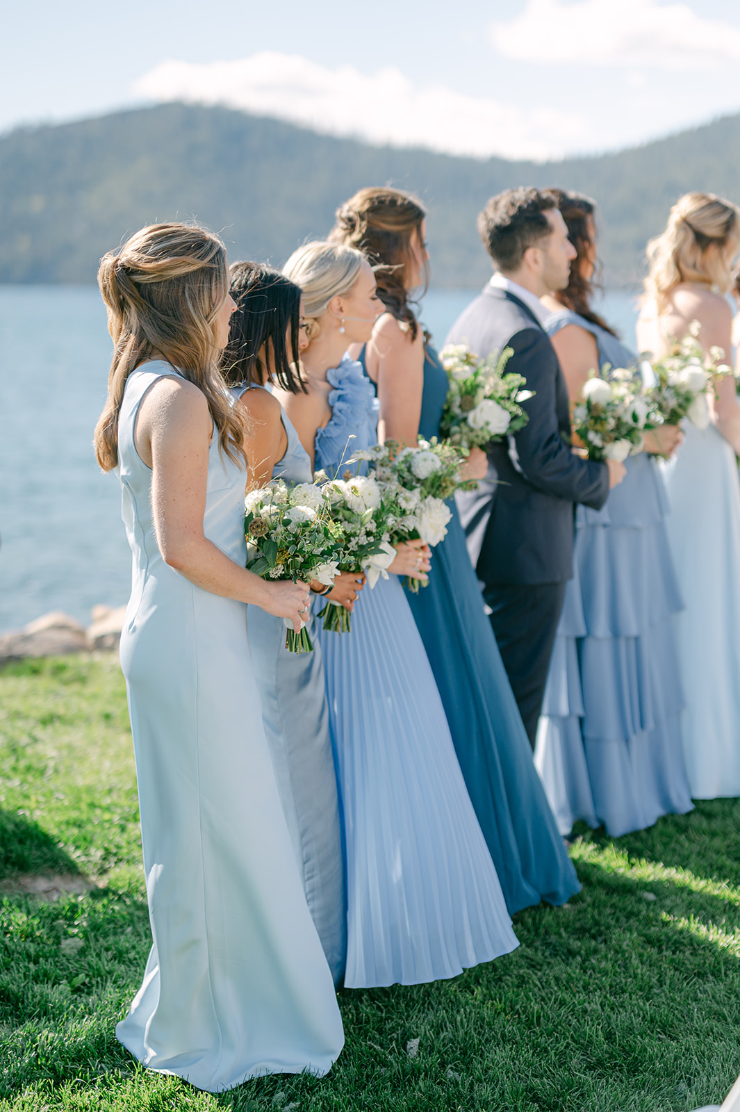 Bridesmaids in mismatched blue dresses standing along the aisle at a lakeside wedding.